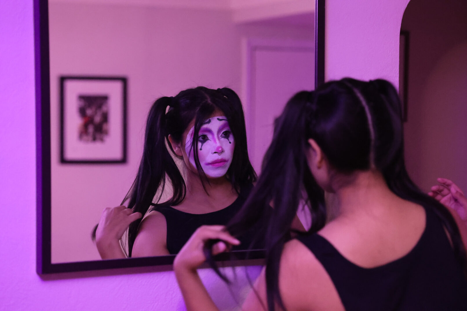 A woman with pigtails playing with her hair while looking in the mirror with her face painted like a clown